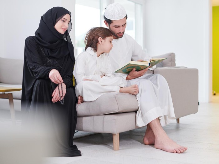 Traditional muslim family parents with children reading Quran and praying together on the sofa before iftar dinner during a ramadan feast at home