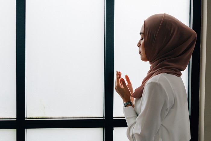 Southeast Asian Islamic female in hijab clasping hands and looking up while standing near window during prayer
