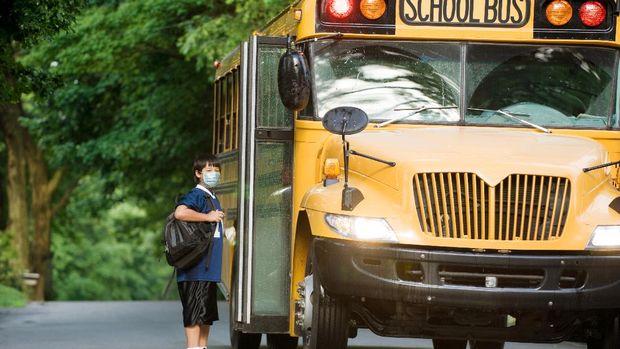 Young female bus driver driving he school bus, wearing a face mask. She is taking care that kids sit down.