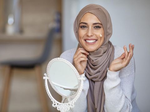 Muslim woman spreading cream over her face while looking in the mirror. Beauty treatment. Female putting on moisturizer on her facial. Muslim woman applying cream to face and looking to mirror