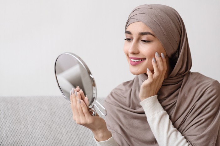 Beauty routine. Attractive muslim girl in hijab holding mirror and looking at her face with perfect soft skin, sitting on sofa at home, free space