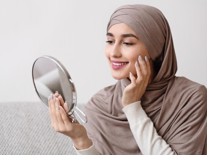 Ilustrasi skincare Ramadhan Beauty routine. Attractive muslim girl in hijab holding mirror and looking at her face with perfect soft skin, sitting on sofa at home, free space