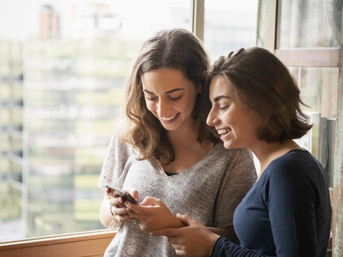 1068831124 Two sisters laughing and looking at messages on cell phone, online, social media, texting