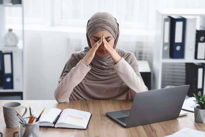 Exhausted arabic businesswoman having migraine after long time work in office, sitting at desk and touching her nose bridge, having bad day