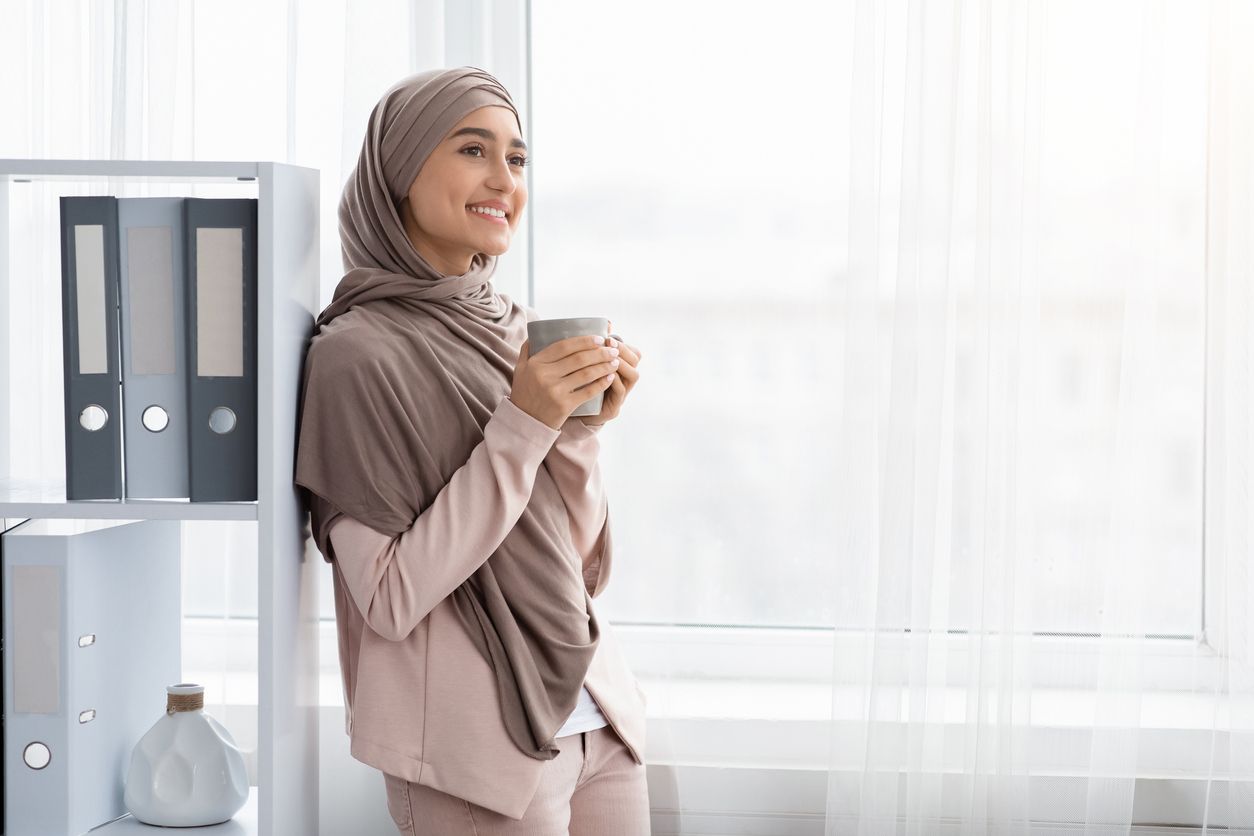 Coffee Break. Cheerful Arabic Businesswoman In Hijab With Cup Of Hot Drink Standing Near Window In Office, Empty Space
