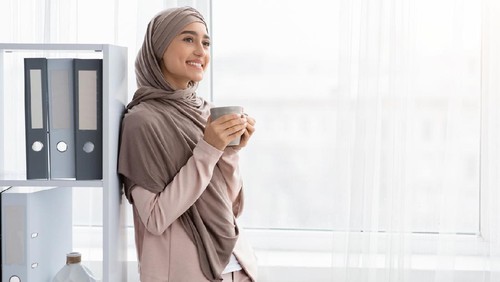 Coffee Break. Cheerful Arabic Businesswoman In Hijab With Cup Of Hot Drink Standing Near Window In Office, Empty Space