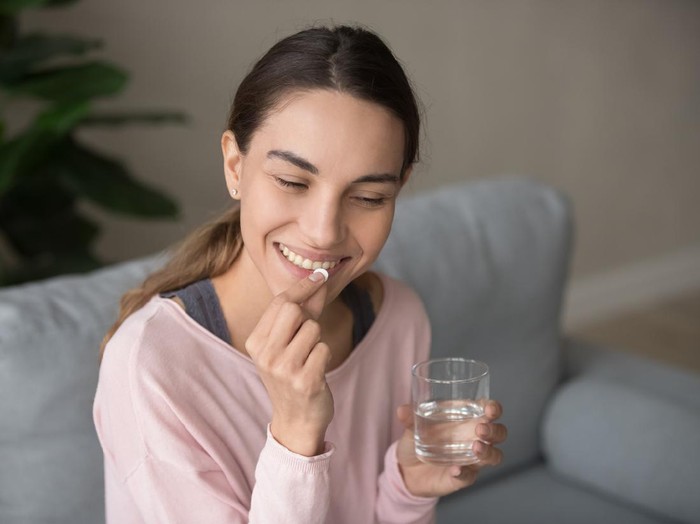 Young happy smiling mixed race woman sitting on a sofa at living room, holding glass of pure water, taking daily vitamin d, c complex, supplements for hair, skin and nail strengthen, healthcare concept.