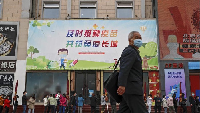 A man wearing a face mask to help curb the spread of the coronavirus walks by masked residents lining up for COVID-19 vaccine at a vaccination site with a board displaying the slogan, 
