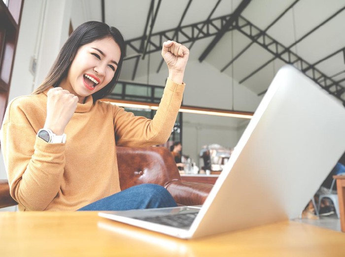 Young attractive asian woman looking at laptop computer feeling happy cheerful or excited expression success or win sitting on desk table in cafe coffee shop in wide close up view with soft tone.
