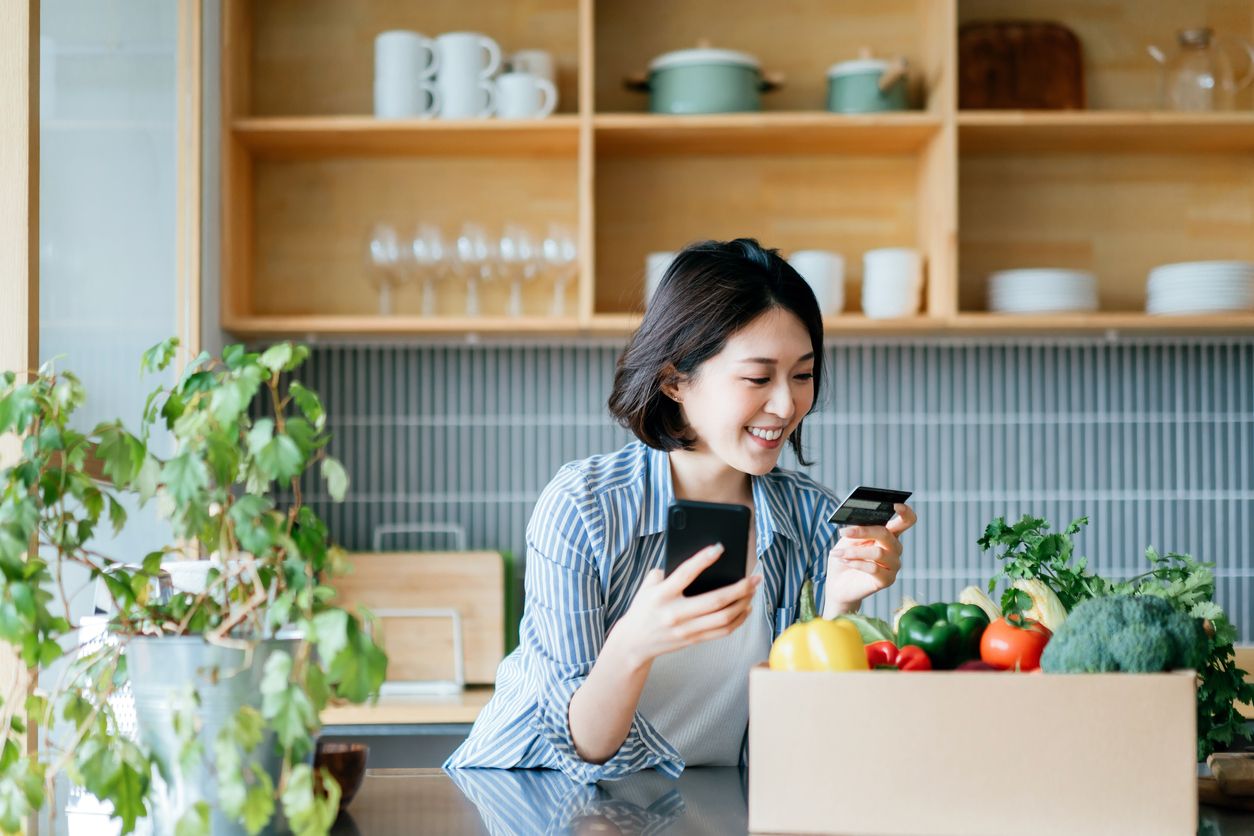 Beautiful smiling young Asian woman grocery shopping online with mobile app device on smartphone and making online payment with her credit card, with a box of colourful and fresh organic groceries on the kitchen counter at home