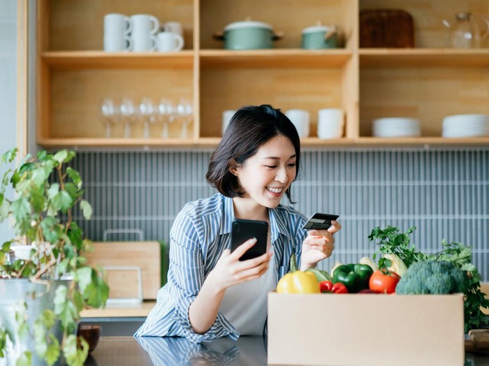 1272443174 Beautiful smiling young Asian woman grocery shopping online with mobile app device on smartphone and making online payment with her credit card, with a box of colourful and fresh organic groceries on the kitchen counter at home