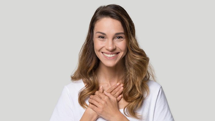 Head shot portrait grateful hopeful young woman holding hands on chest, pleased young female looking at camera, feeling love, gratitude, appreciation, thanking fate, isolated on studio background