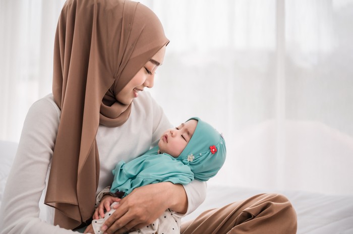 Happy muslim mother holding adorable little baby daughter wearing hijab in her arms on white bed in bedroom. Arab young mom wearing a head scarf, and shes sitting on the bed while holding her baby.