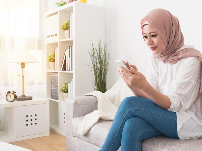 elegant attractive female muslim using mobile smartphone searching online information when she sitting on sofa couch relaxing.