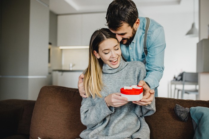 Young couple is at home. They are sitting on a couch, head to head and holding a present