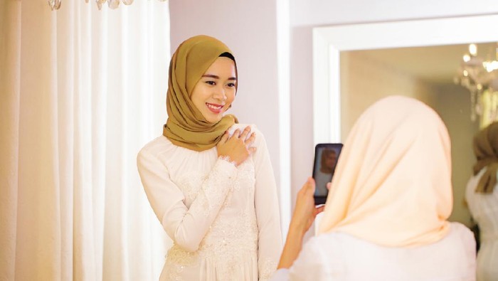 Young Muslim woman photographing her friend trying on a wedding dress in the bridal shops fitting room.