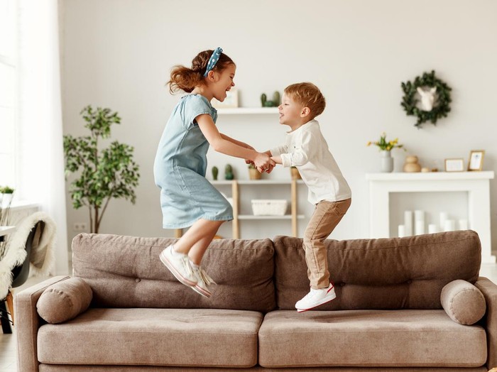 Side view of cheerful boy and girl holding hands and jumping on couch while having fun at home together