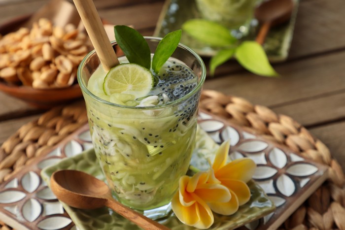 Es Kuwut, a Balinese cold dessert drink of honeydew melon, coconut, basil seeds, and lime. The drink is served in a clear tall glass and garnished with lime half, lime leaves, and a bamboo straw. The glass is placed on a ceramic saucer embossed with frangipani flower. Fresh frangipani flowers and a wooden spoon are also placed on the saucer. The saucer is then arranged on large wooden coaster carved with flowery shape; together with local snack of roasted peanuts. The image was shot outdoor on the terrace with natural lighting.
