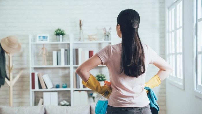 back view of asian housekeeper looking at the clean living room after she tidied up. young wife finished house chores putting hands in waist watching the bookshelf beside the sunlight window.