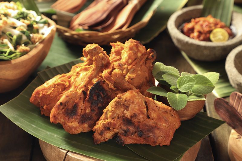 Ayam Bakar Padang, the traditional spicy grilled chicken from Minangkabau cuisine, West Sumatra. The chicken pieces are arranged on a wooden cutting block lined with banana leaf. Accompanied with red chili sauce and a vegetable salad.