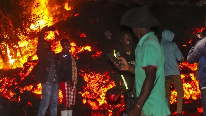 Saat Warga Kongo Asik Selfie di Depan Lava Gunung Berapi Residents check the damages caused by lava from the overnight eruption of Mount Nyiragongo, seen in background, in Buhene, on the outskirts of Goma, Congo in the early hours of Sunday, May 23, 2021. Congo's Mount Nyiragongo erupted for the first time in nearly two decades Saturday, turning the night sky a fiery red and sending lava onto a major highway as panicked residents tried to flee Goma, a city of nearly 2 million. (AP Photo/Justin Kabumba)