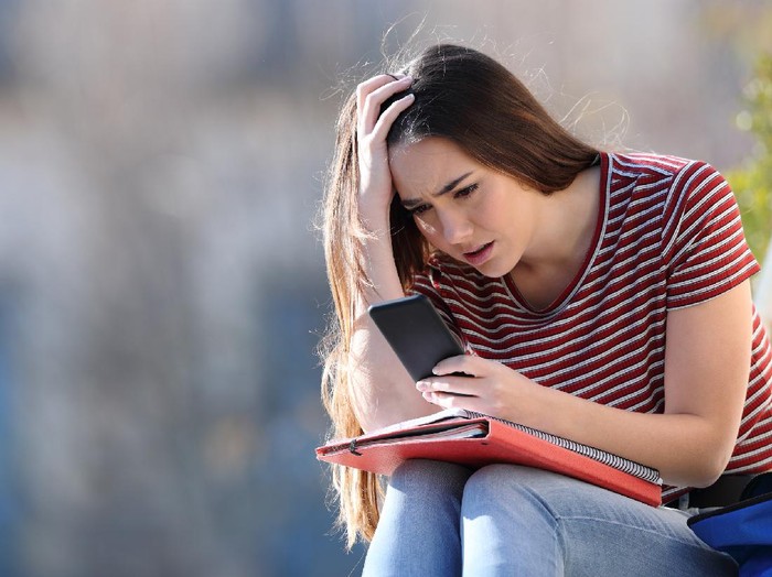 1097258096 A young blonde woman grimaces, hand to her face as she hears something on her smart phone, obviously bad news.