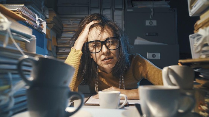 Stressed exhausted woman sitting at office desk and working overtime, she is overloaded with work
