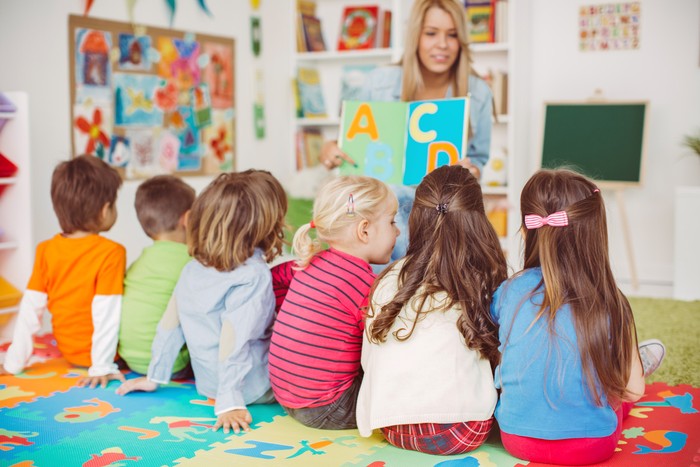 Beautiful teacher and group of toddlers sitting on the floor drawing using paper and pencil around lots of toys at kindergarten