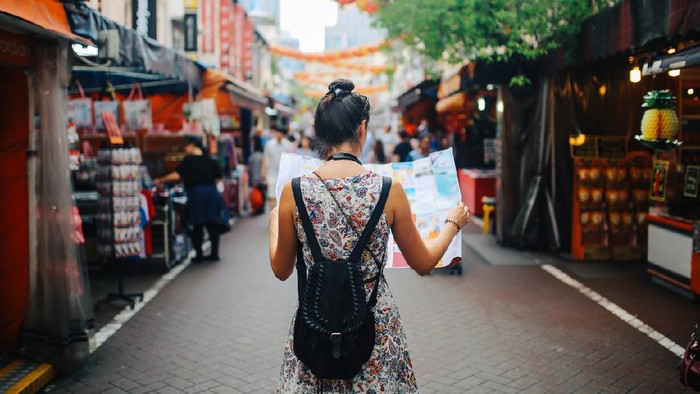 Rear view image of a young brunette woman. She is enjoying the walk and exploring the city, wearing a casual but fashionable dress, sightseeing and shopping on the Singapore street market. She is holding a large city map, checking out where to go next.