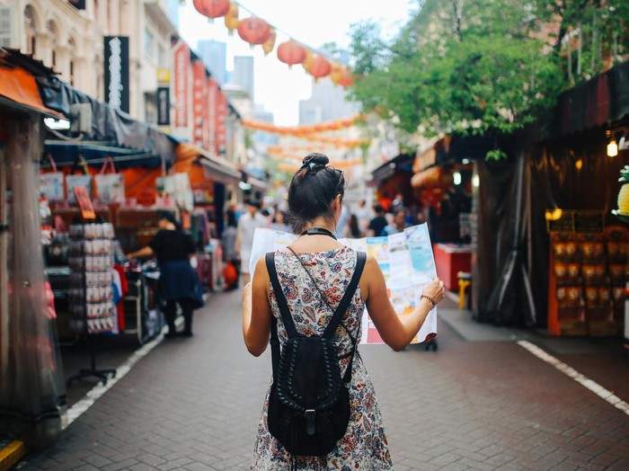 Rear view image of a young brunette woman. She is enjoying the walk and exploring the city, wearing a casual but fashionable dress, sightseeing and shopping on the Singapore street market. She is holding a large city map, checking out where to go next.