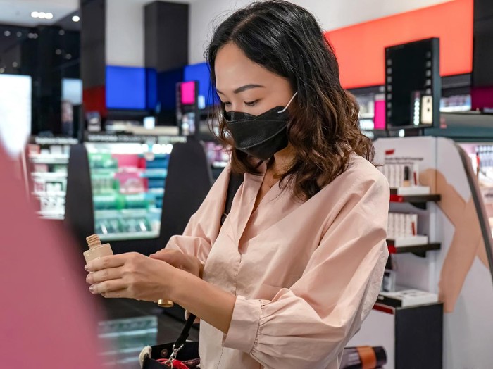 1284175588 Portrait shot of a Southeast Asian woman with a face mask applying liquid foundation to her hand to match its color with her skin tone, during her make up shopping amidst Covid 19 pandemic