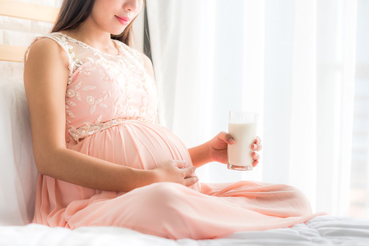 Happy pregnant woman holding a glass of fresh milk and touching her belly with care by the window in bedroom