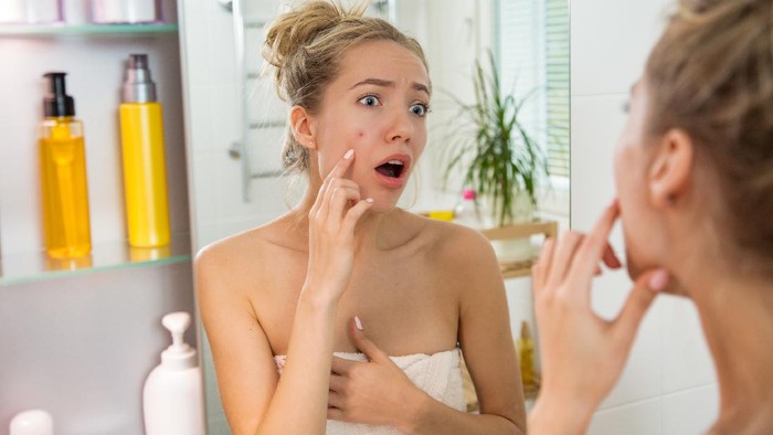 Young beautiful woman surprised to see pimple on skin. Unhappy girl with stunned funny face standing in towel in bathroom, looking in the mirror, pointing to zit