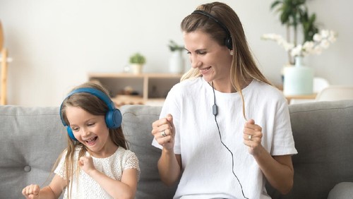 Happy young mother and funny child daughter having fun laughing dancing sitting on couch listening to music in earphones together, smiling mom and kid wearing headphones enjoy favorite songs at home