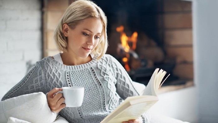 A beautiful young woman reading a book and enjoying a warm beverage near a fireplace