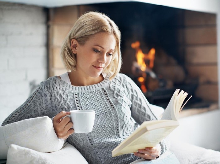 Ilustrasi Baca Buku A beautiful young woman reading a book and enjoying a warm beverage near a fireplace