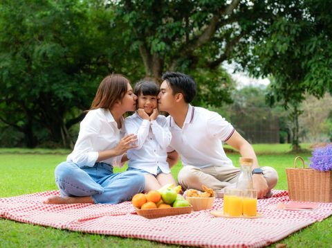 Ilustrasi Piknik di Halaman Asian teen family happy holiday picnic moment in the park with father, mother kissing daughter looking at camera and smile to happy spend vacation time together in green garden with fruit and food.
