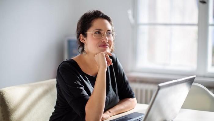 Businesswoman sitting in front of a laptop and thinking. Female is sitting at boardroom table with her laptop.
