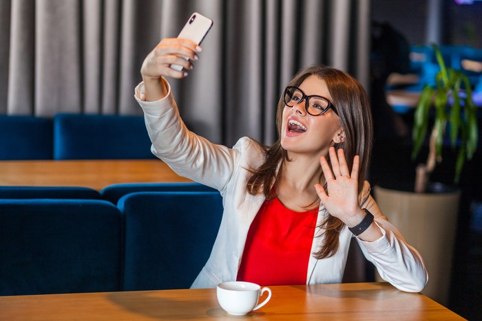 Portrait of happy joyful beautiful stylish brunette young woman in glasses sitting on video call or doing selfie with toothy smile and happiness waving hand. indoor studio shot, cafe office background