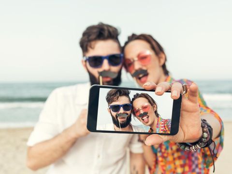 Cheerful hipster couple making selfies on a cell phone with fake paper moustaches on a sunny summer day at a secluded beach