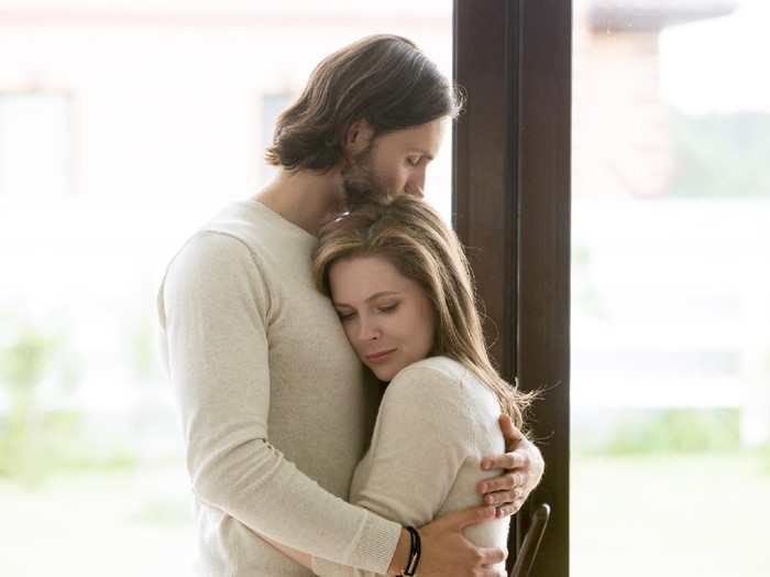 1055880362 Sad young married couple embracing standing in living room opposite window at home. Sorrowful wife and husband feels unhappy, thinking about problems in relations, miscarriage, misbirth or infertility