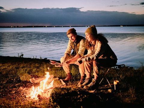 Positive young couple sitting at lake shore and making sausages on fire during night camping