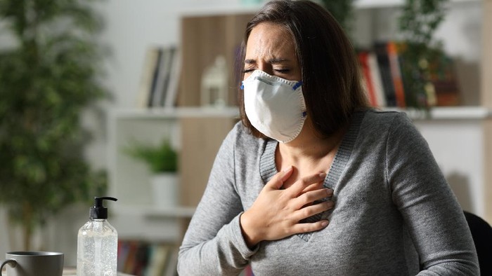 1221585409 Woman with protective mask suffocating due coronavirus holding chest sitting on a desk at nigh at home