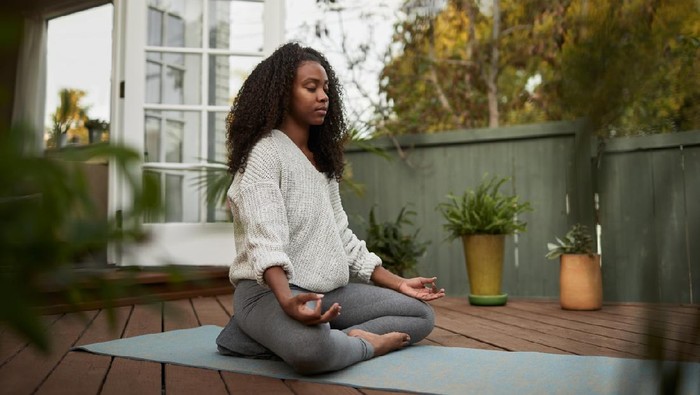 Young African American woman sitting on exercise mat outside on her patio and meditating in the lotus pose during a yoga session