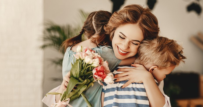 Happy Mothers Day! Children boy and girl congratulate smiling mother, hugs her  and give her flowers   bouquet of tulips during holiday celebration in kitchen at home