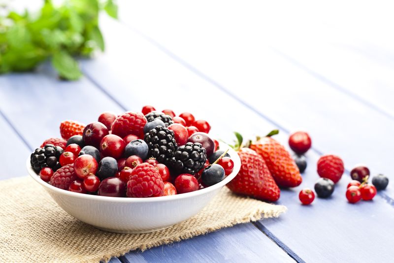 White bowl filled with fresh mixed organic berries on blue garden table. Some berries are scattered outside the bowl. DSRL studio photo taken with Canon EOS 5D Mk II and Canon EF 100mm f/2.8L Macro IS USM