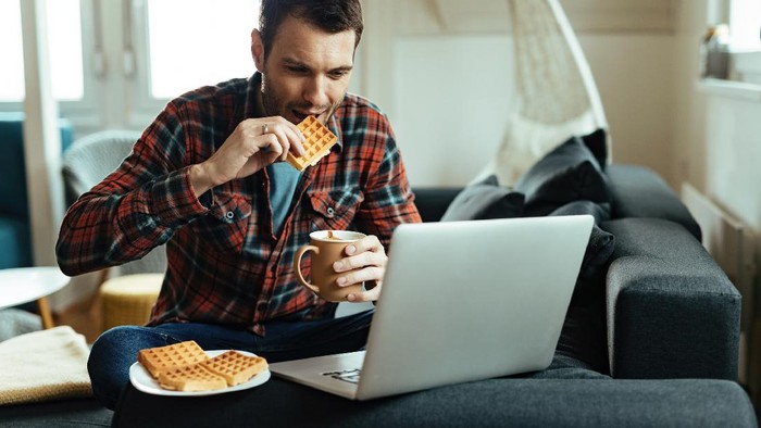 Smiling man using laptop while drinking coffee and eating waffle in the living room.