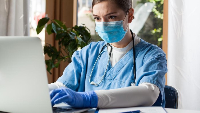 Young caucasian female doctor working on laptop computer in office,wearing protective glove and face mask,Electronic Medical Records EMR concept,remote tele health medicine during Coronavirus pandemic