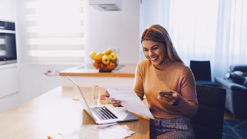 Cute caucasian smiling blonde woman in sweater holding bills in one hand and in other smart phone. On table are laptop and bills. Apartment interior.