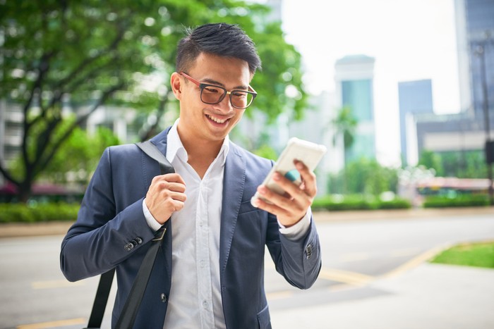 Young malaysian businessman in the streets of downtown Kuala Lumpur smiling while looking to his his smart phone for a crowed sourced taxi to arrive . Kuala Lumpur, Malaysia, Asia